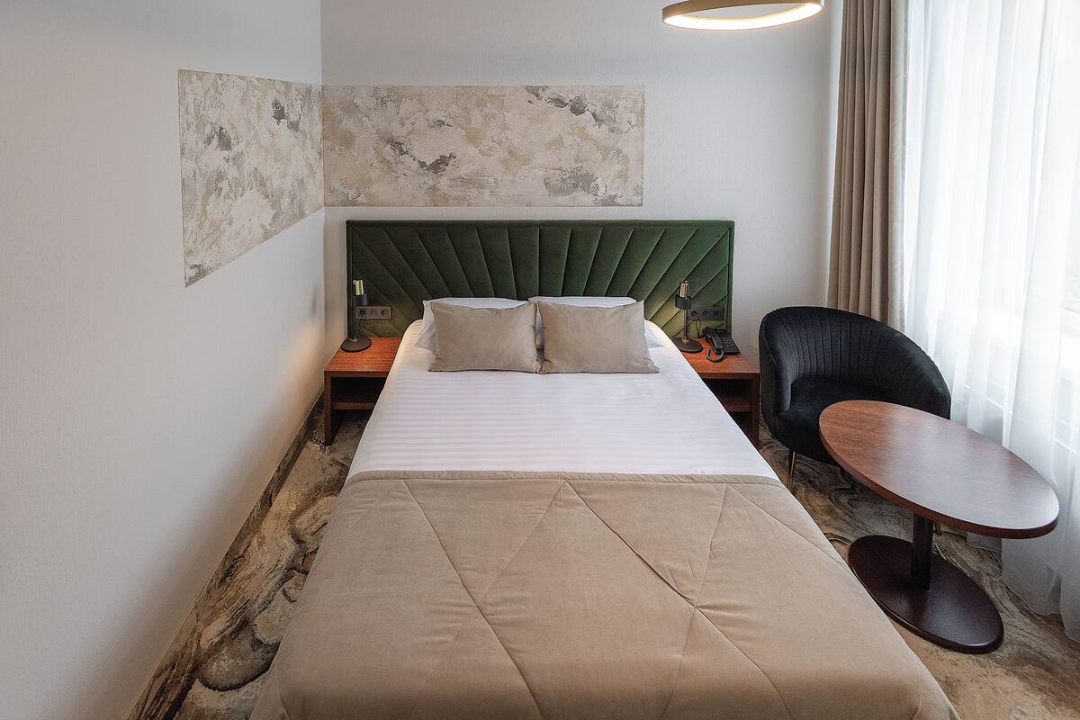 A hotel room with a bed, a green upholstered headboard, a round wooden table, a black chair, and a large abstract painting on the wall.
