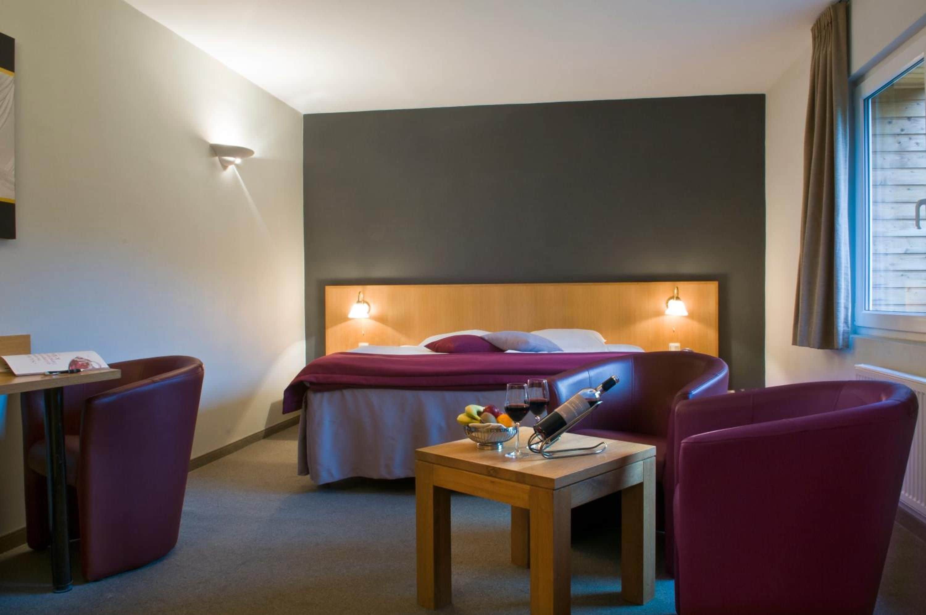 A hotel room with a bed, a desk, a chair, and a table with a bottle and glasses.