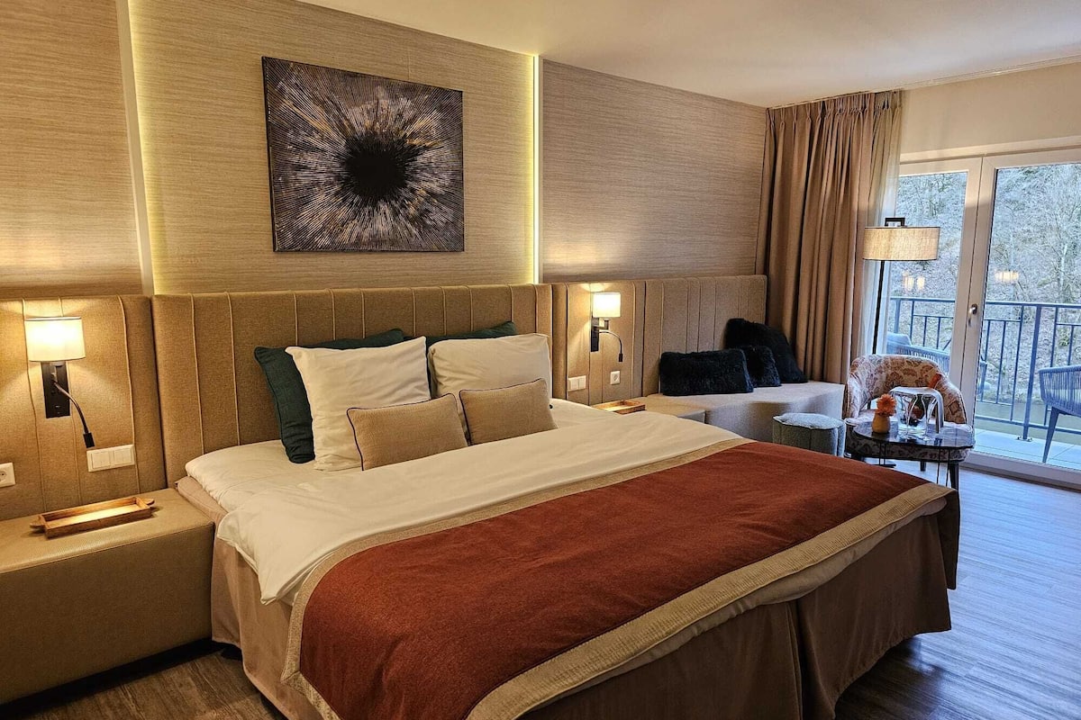 Quarto de hotel com uma cama grande, duas mesas de cabeceira, um sofá, uma pequena mesa com abajur e vista para árvores pela janela.