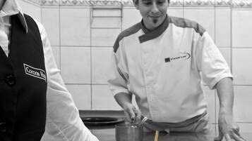 Un chef, vêtu d’un uniforme blanc et d’un gilet noir, présente un plat sur une assiette blanche, composé de légumes et d’une brochette de viande.