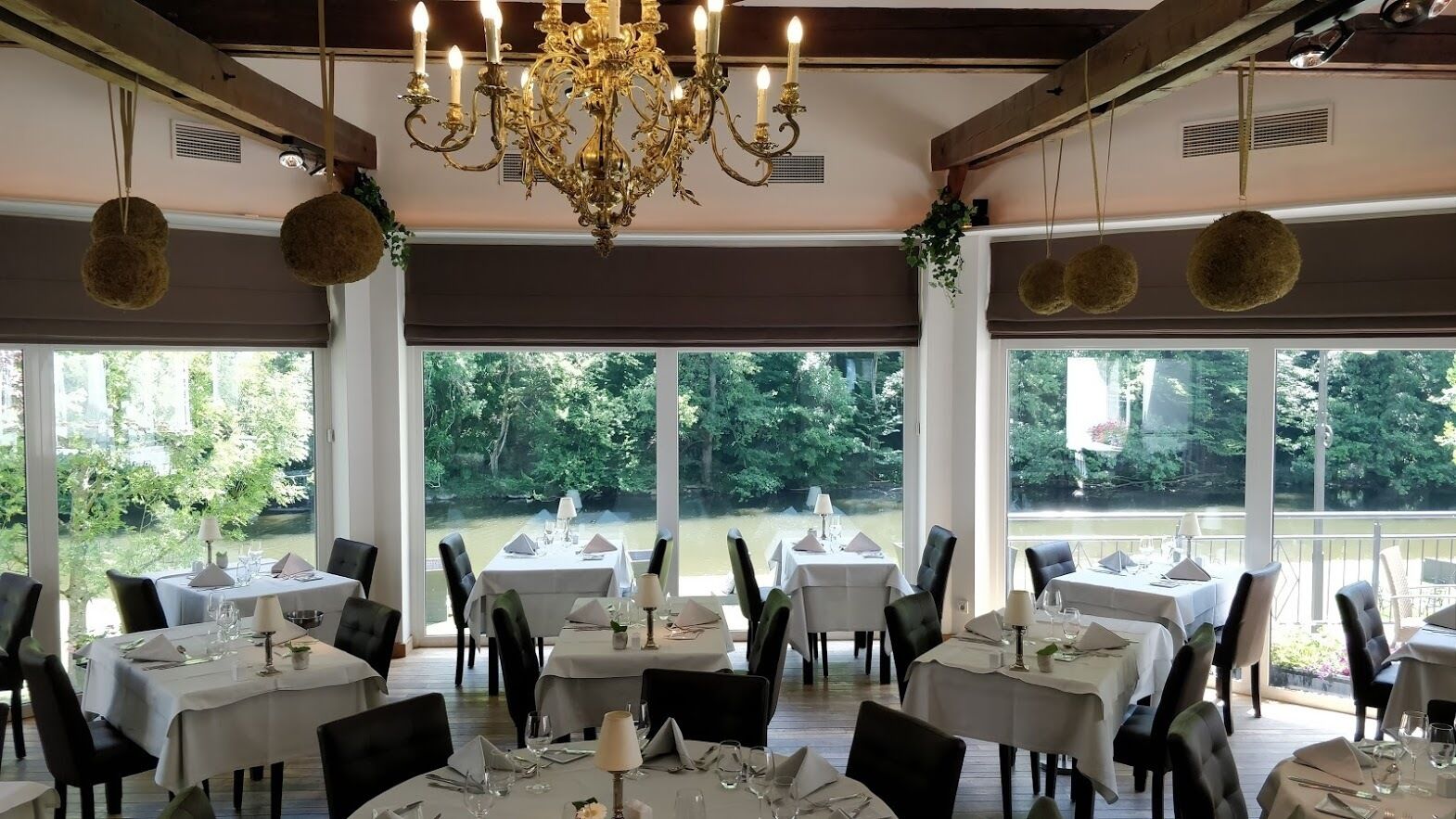 A restaurant with tables set for dining, large windows with a view of greenery, and a chandelier.