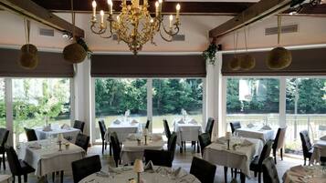 A restaurant with tables set for dining, large windows with a view of greenery, and a chandelier.