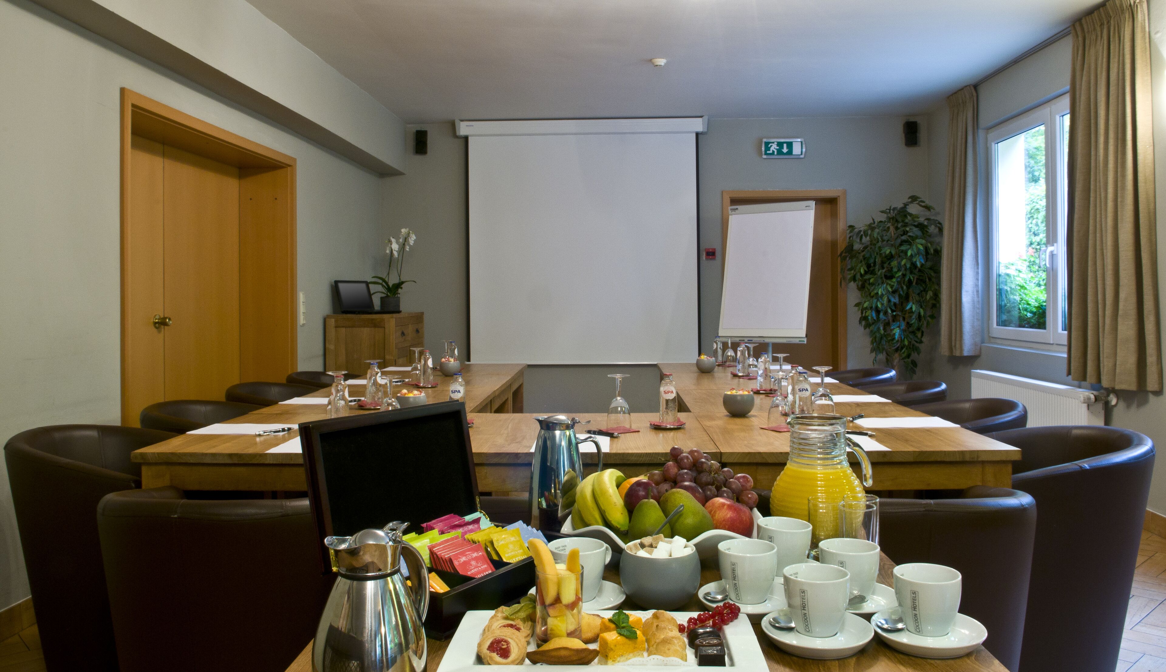 Un salón de conferencias con una larga mesa dispuesta para una reunión, equipado con una pantalla de proyector, una pizarra y una planta.