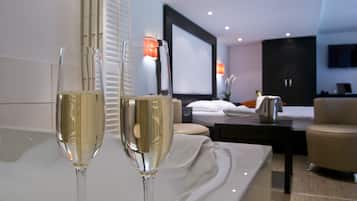 Quarto de hotel moderno com uma cama, uma mesa de cabeceira com abajur e duas taças de champanhe sobre uma mesa branca.