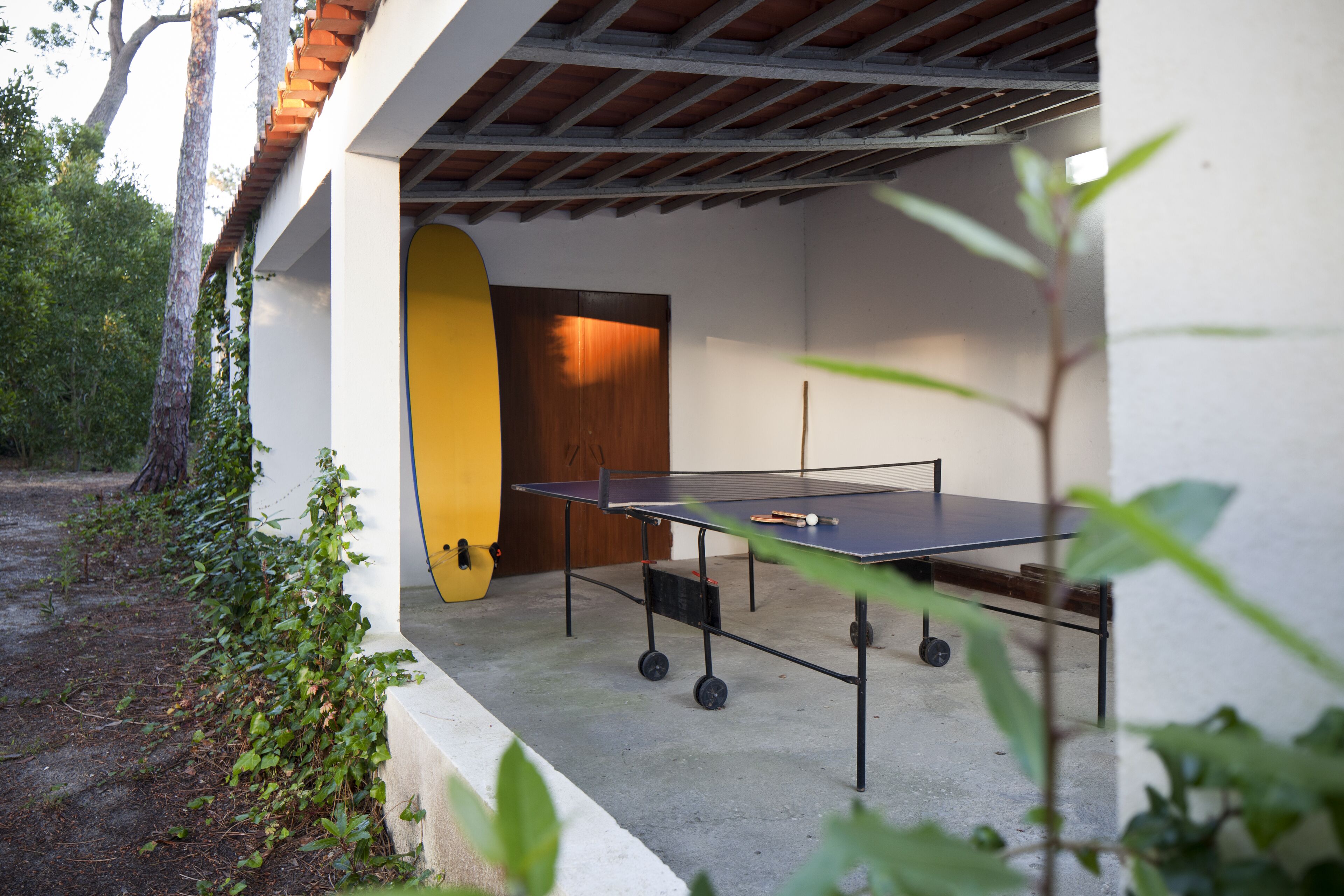 An outdoor ping pong table with a yellow surfboard leaning against a wall, surrounded by greenery.