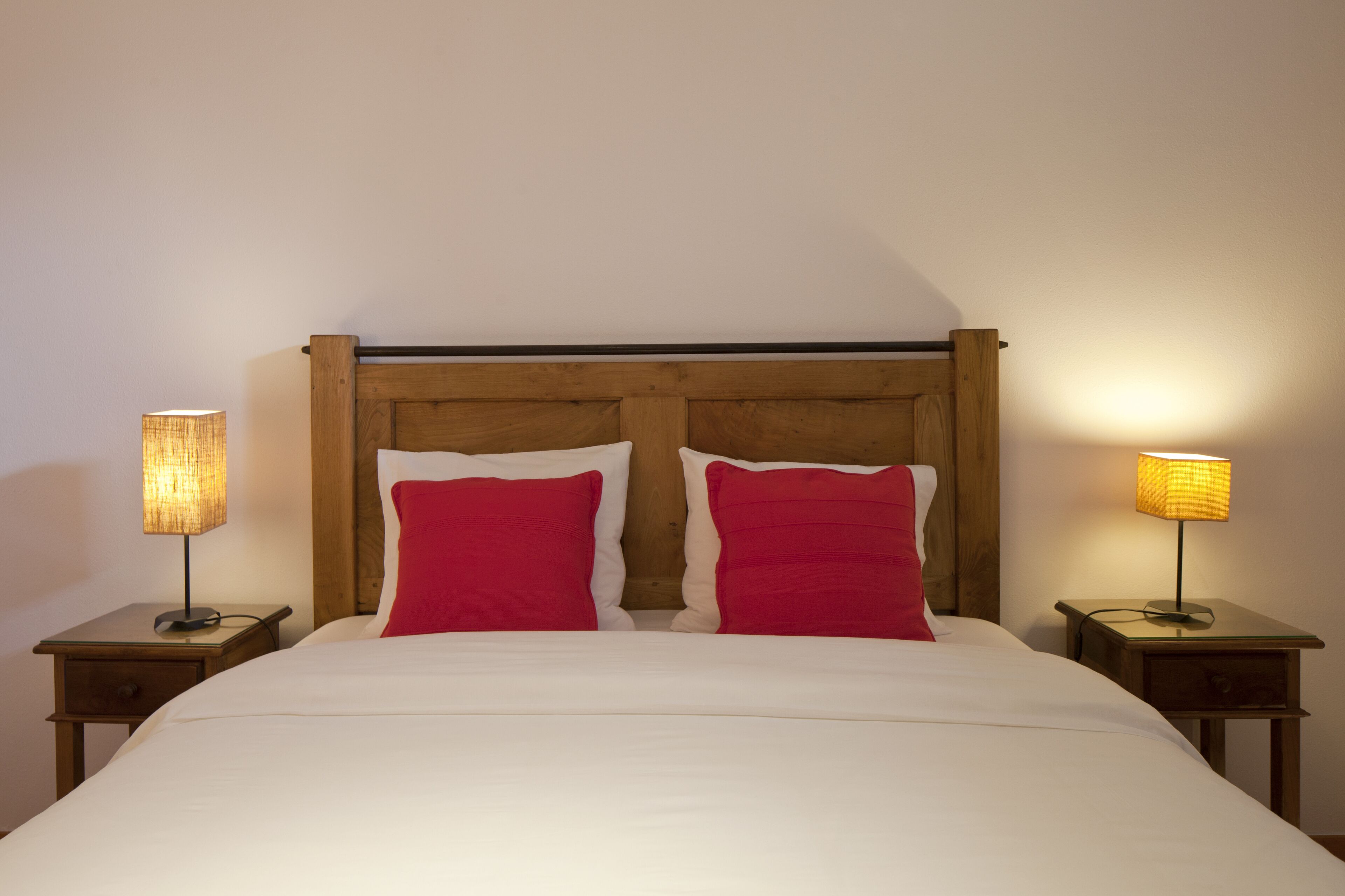 Una cama bien hecha con tablero de madera, dos almohadas rojas y dos lámparas de noche sobre mesitas de madera.
