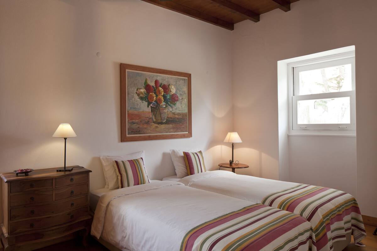 A bedroom with two beds, a wooden dresser, two bedside lamps, a painting of flowers, and a window with natural light.