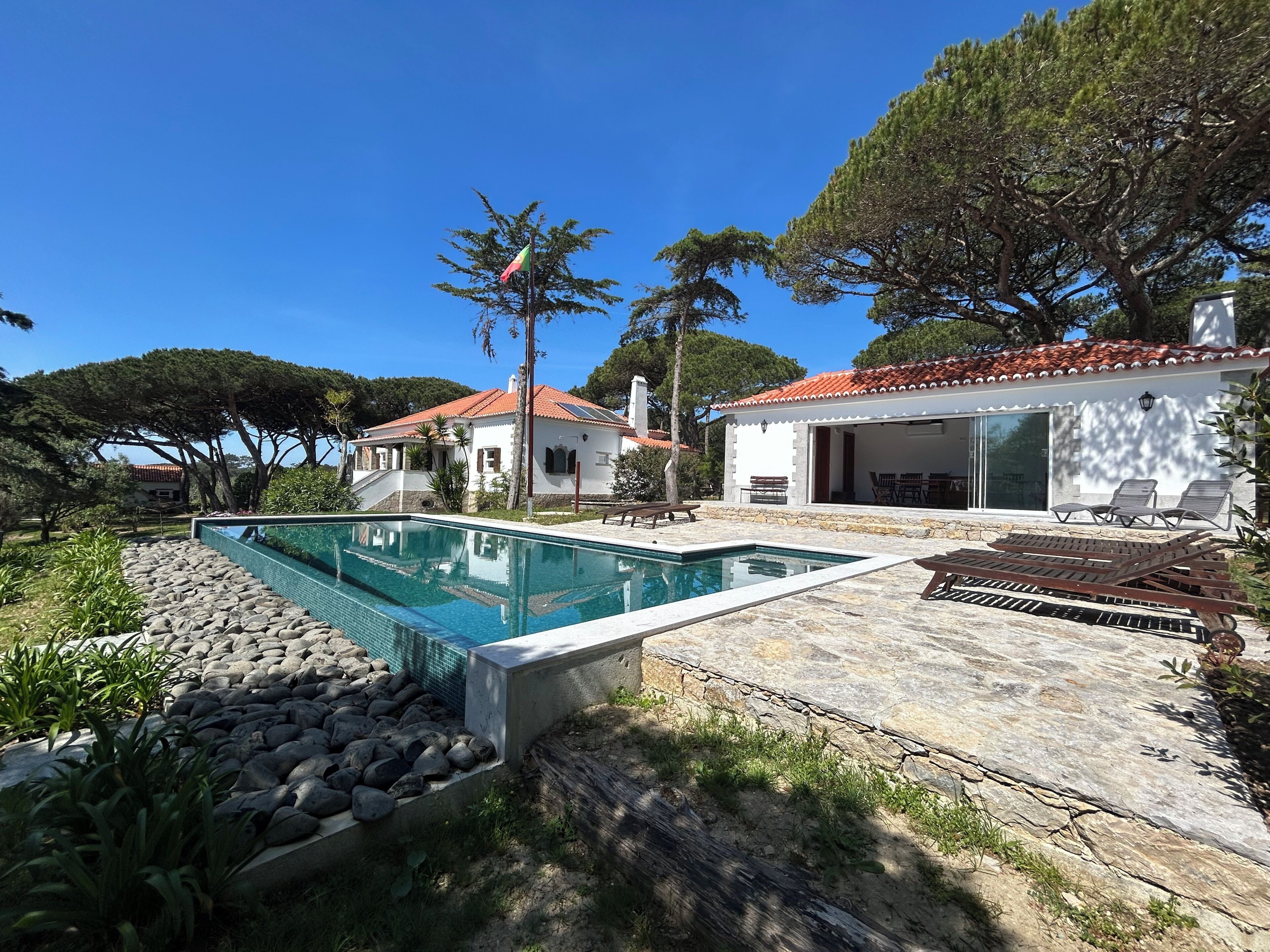 A swimming pool with a stone border, a patio with lounge chairs, and a building with a red-tiled roof.