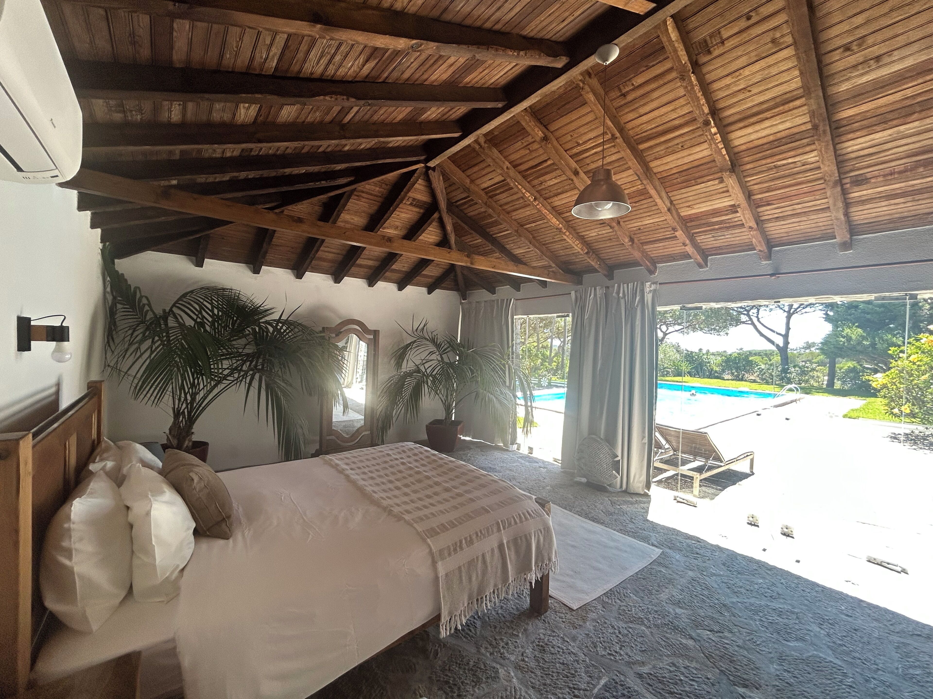 Un dormitorio con una cama grande, vigas de madera en el techo, vistas a una piscina y plantas en macetas.