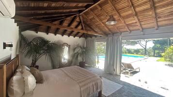 Un dormitorio con una cama grande, vigas de madera en el techo, vistas a una piscina y plantas en macetas.