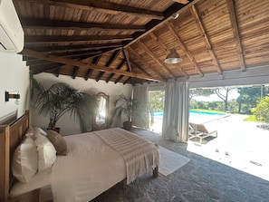 Ein Schlafzimmer mit einem großen Bett, Holzbalkendecke, Blick auf einen Swimmingpool und Blumentöpfen.