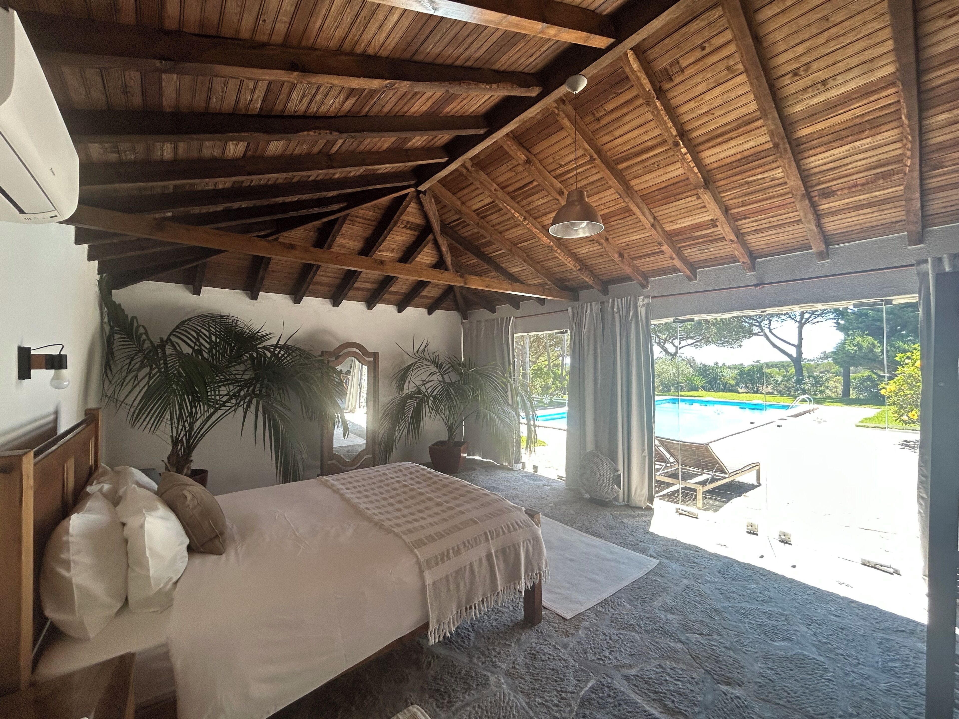 A bedroom with a large bed, wooden ceiling beams, a view of a swimming pool, and potted plants.