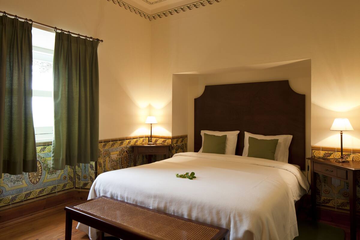 A neatly made bed with a headboard, two bedside lamps, a bench, and a window with curtains.