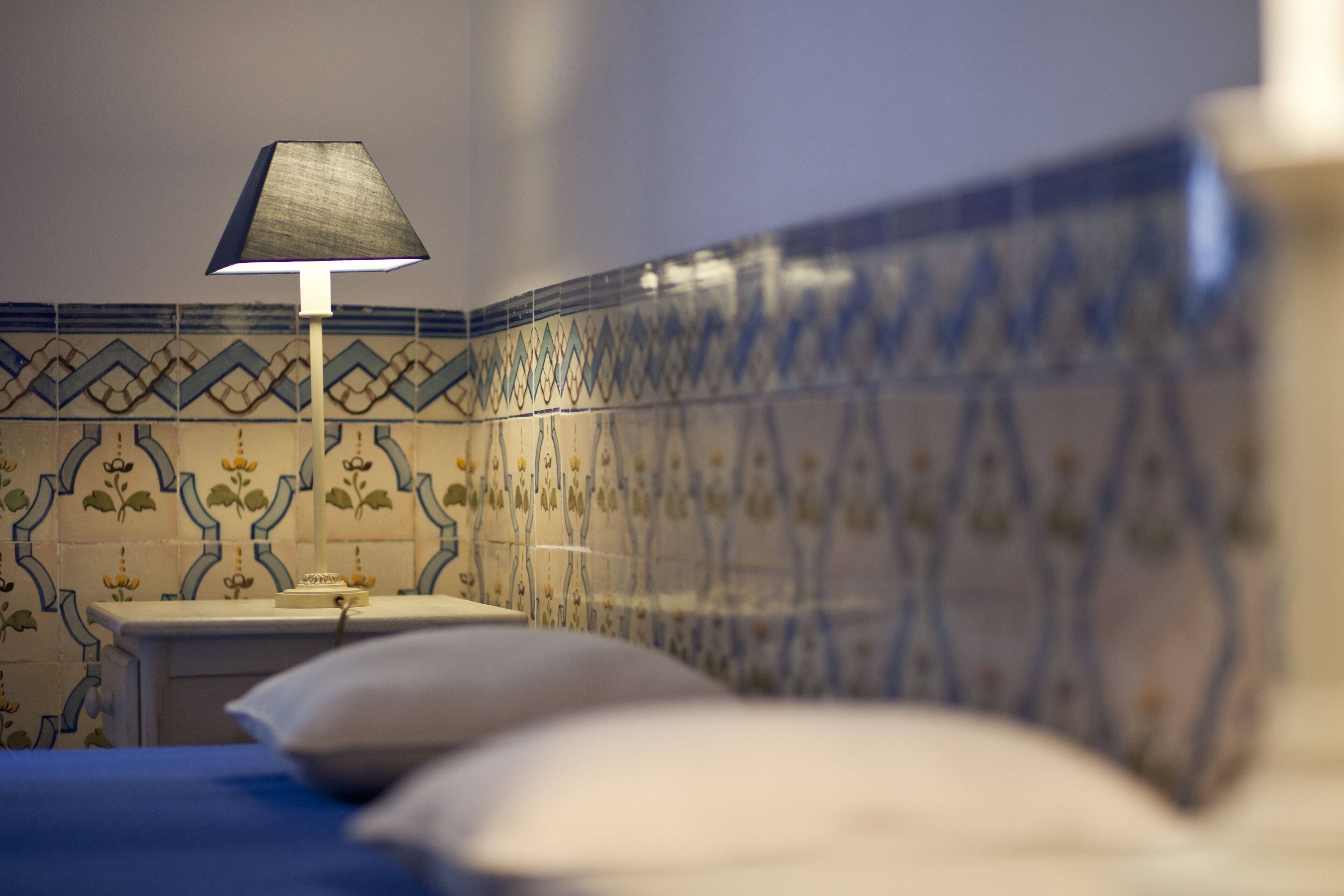 A bed with a white pillow, a bedside table with a lamp, and a wall with blue and white patterned tiles.