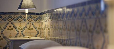 A bed with a white pillow, a bedside table with a lamp, and a wall with blue and white patterned tiles.