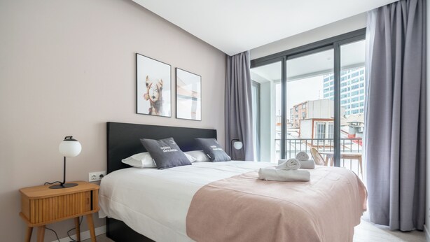 A modern hotel room with a large bed, a bedside table with a lamp, and a view of the city through large windows. -  Limehome Barcelona Rector Triadó (Barcelona)