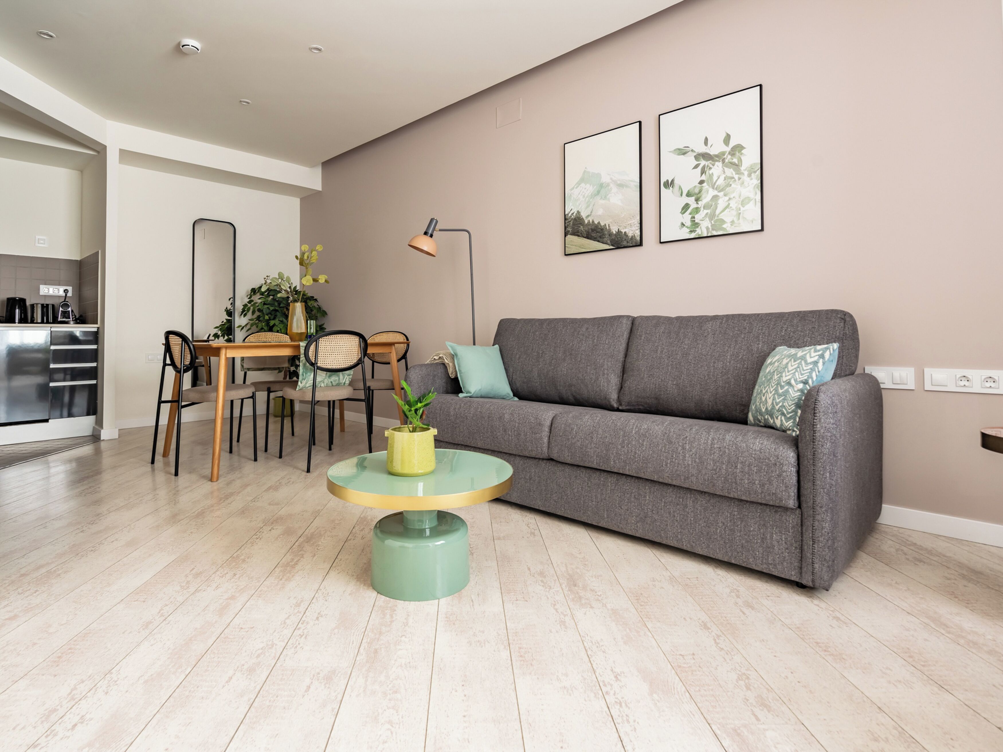 Una sala de estar moderna con un sofá gris, una mesa de centro redonda con una planta, un comedor con una mesa y sillas de madera, y dos cuadros enmarcados en la pared.