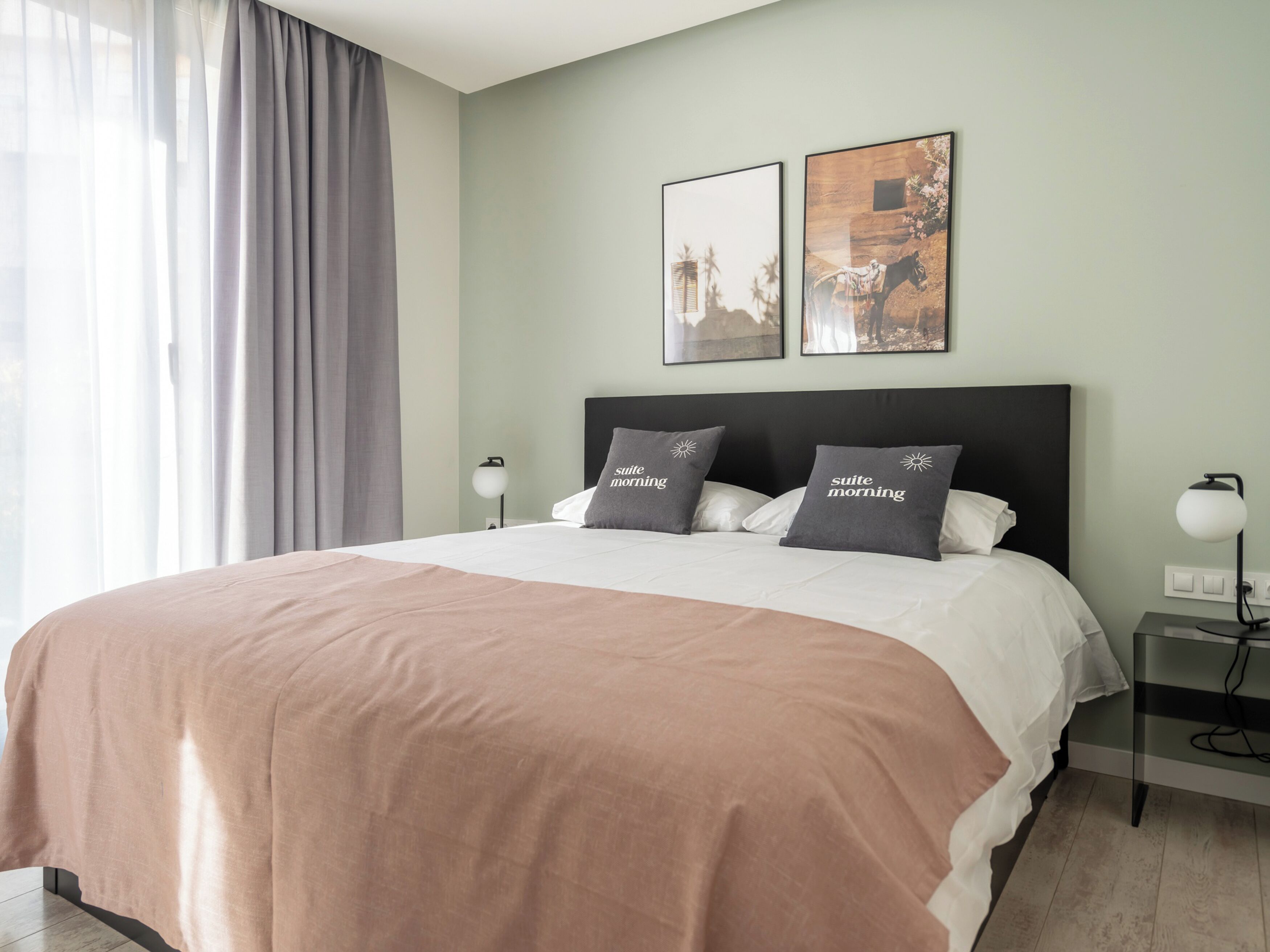 Un lit bien fait, recouvert d’une couvre-lit rose, avec deux oreillers ornés de l’inscription « suite morning », une table de chevet avec une lampe et deux tableaux encadrés accrochés au mur.