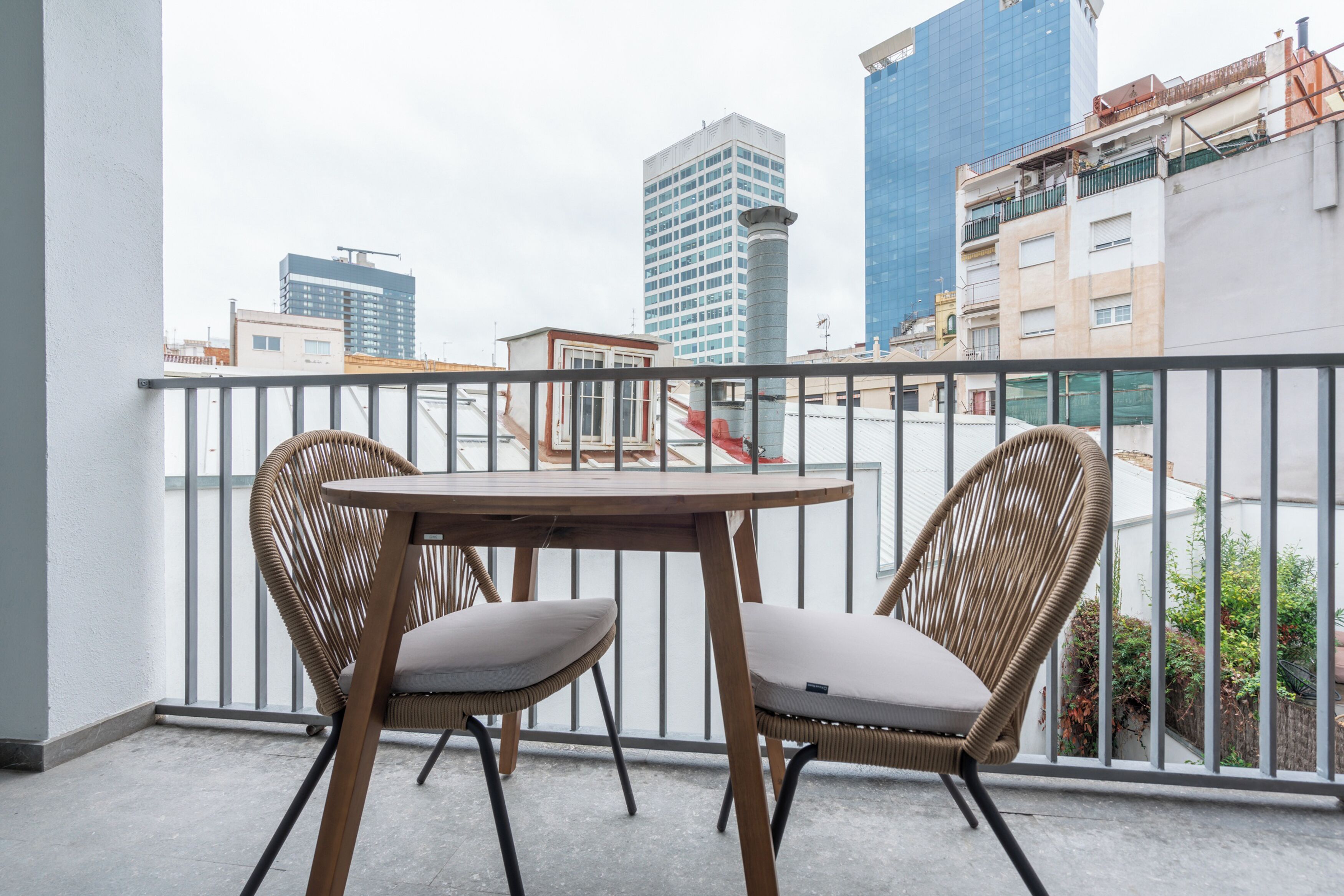 A balcony with two wicker chairs and a small table, overlooking a cityscape with modern buildings.