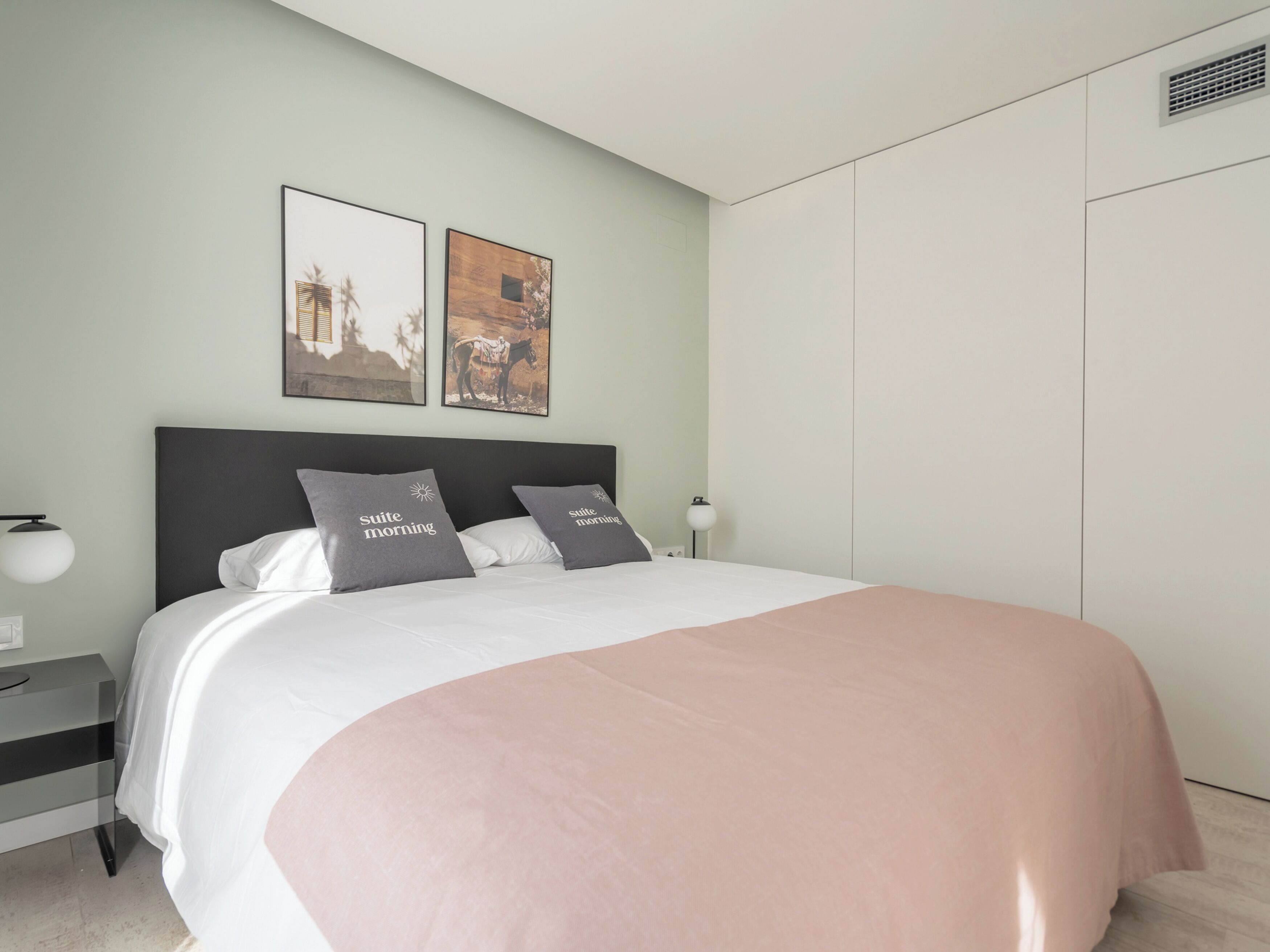 A modern bedroom with a bed, two pillows, a bedside table, a lamp, and two framed pictures on the wall.