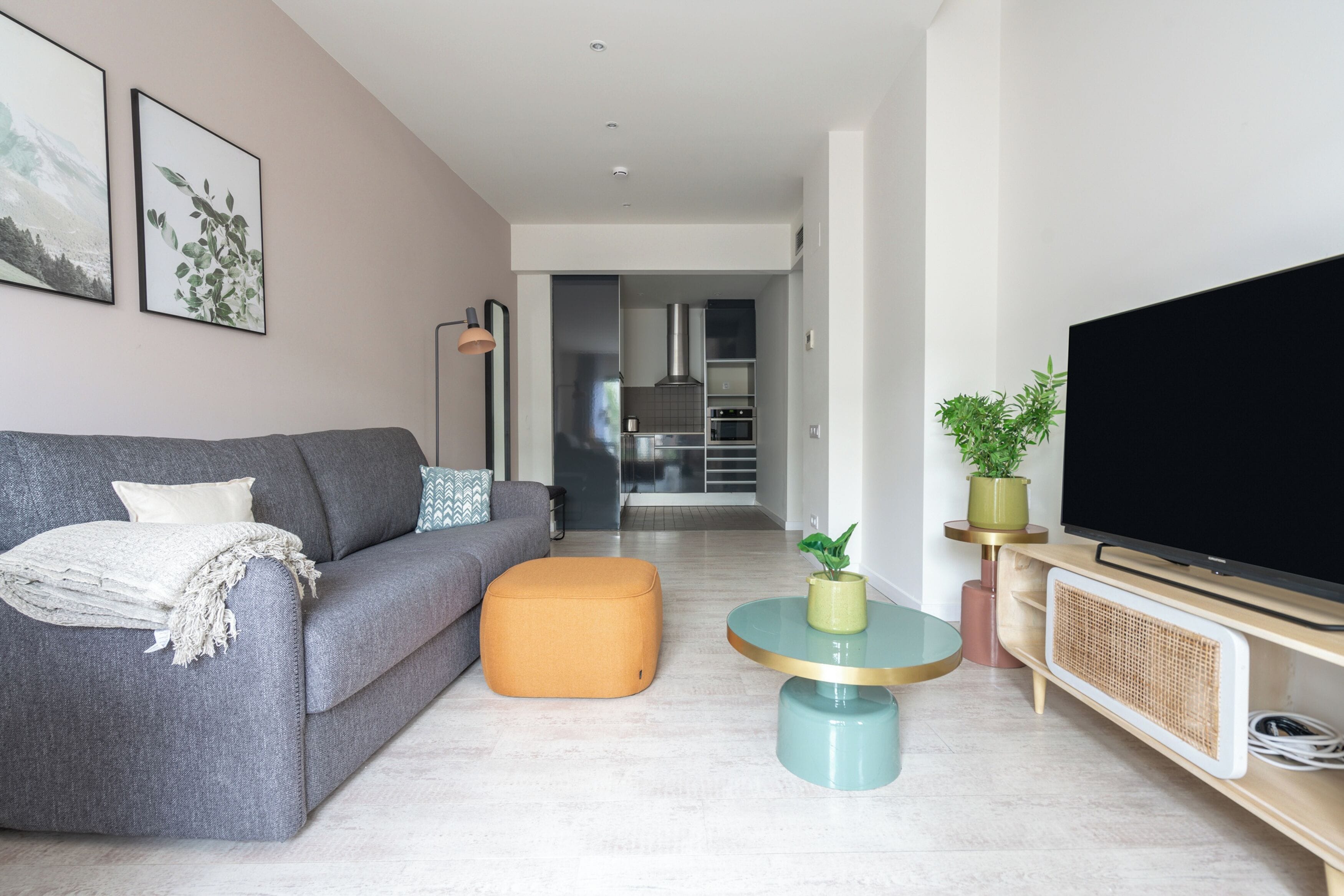 A modern living room with a grey sofa, an orange ottoman, a round table with a plant, and a flat-screen TV on a wooden stand.