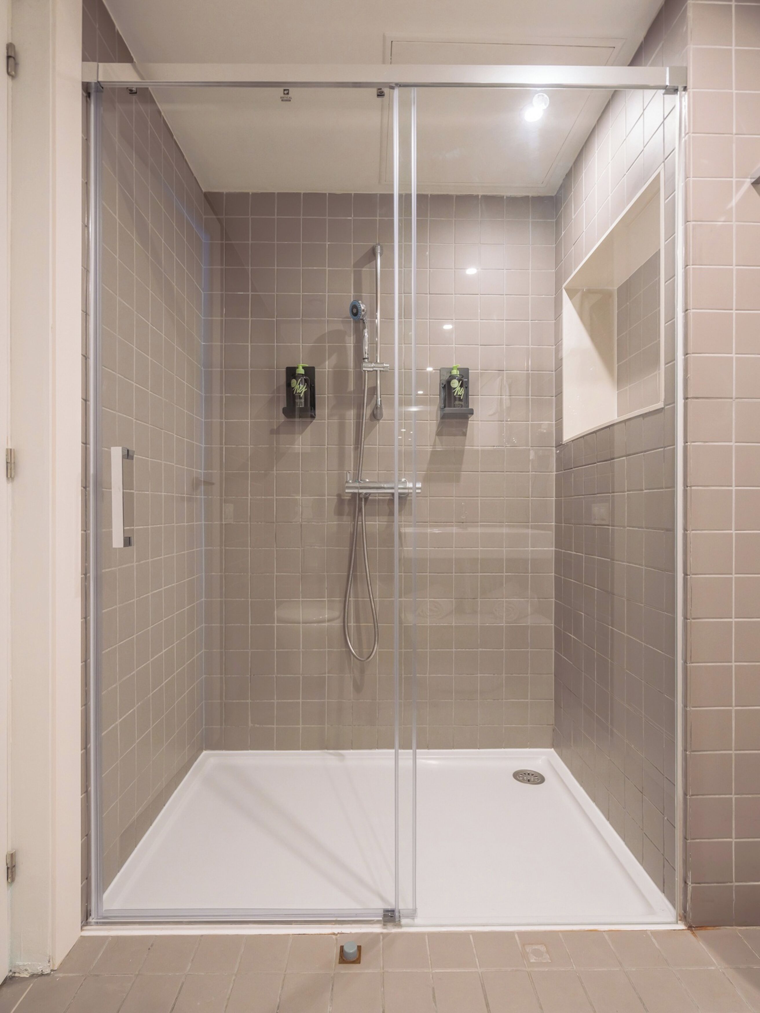 Eine bodenebene Dusche mit Glastür, beige geflieste Wände und einem weißen Boden.