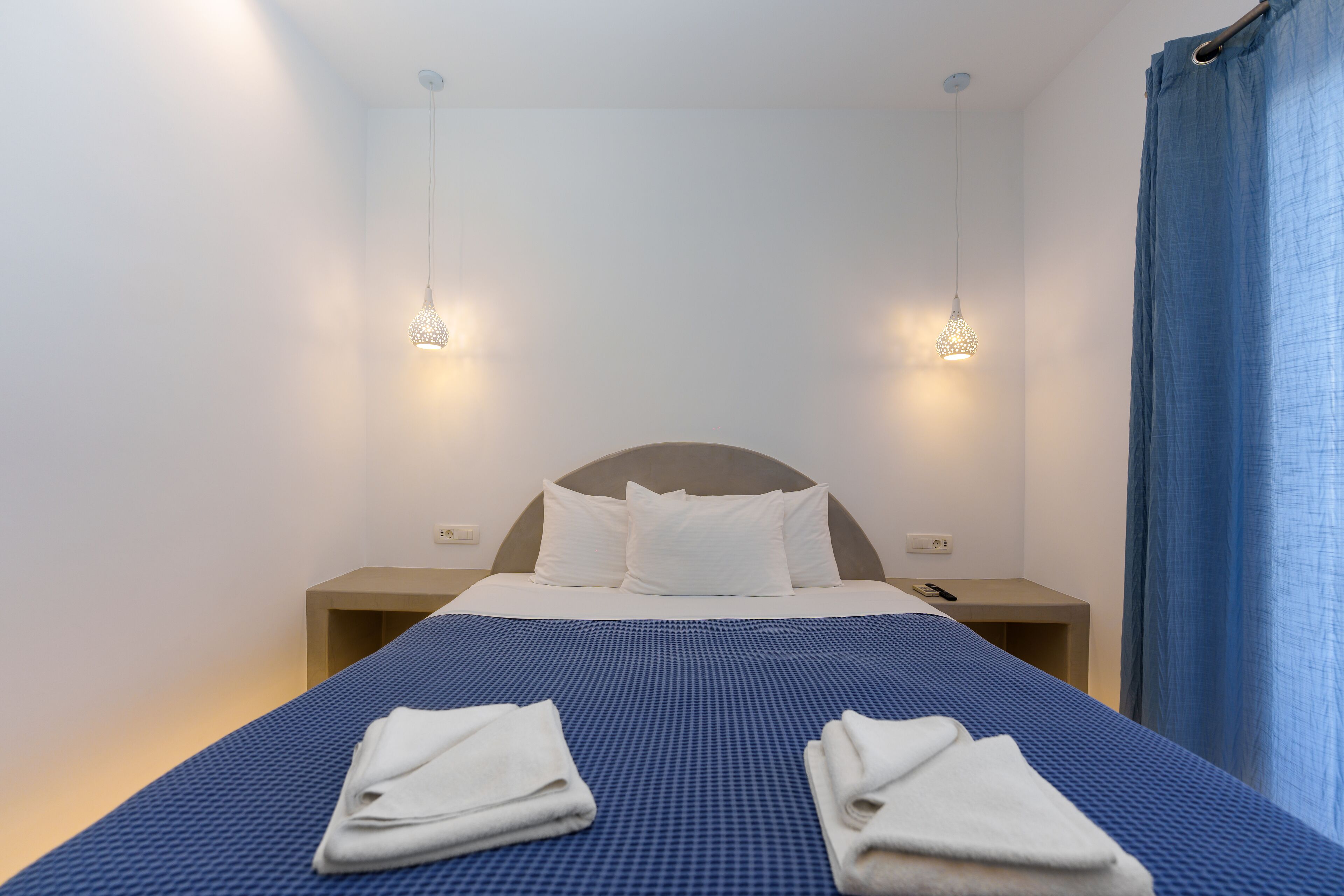 A neatly made bed with a blue bedspread and white pillows, two bedside lamps, and a blue curtain on the right.