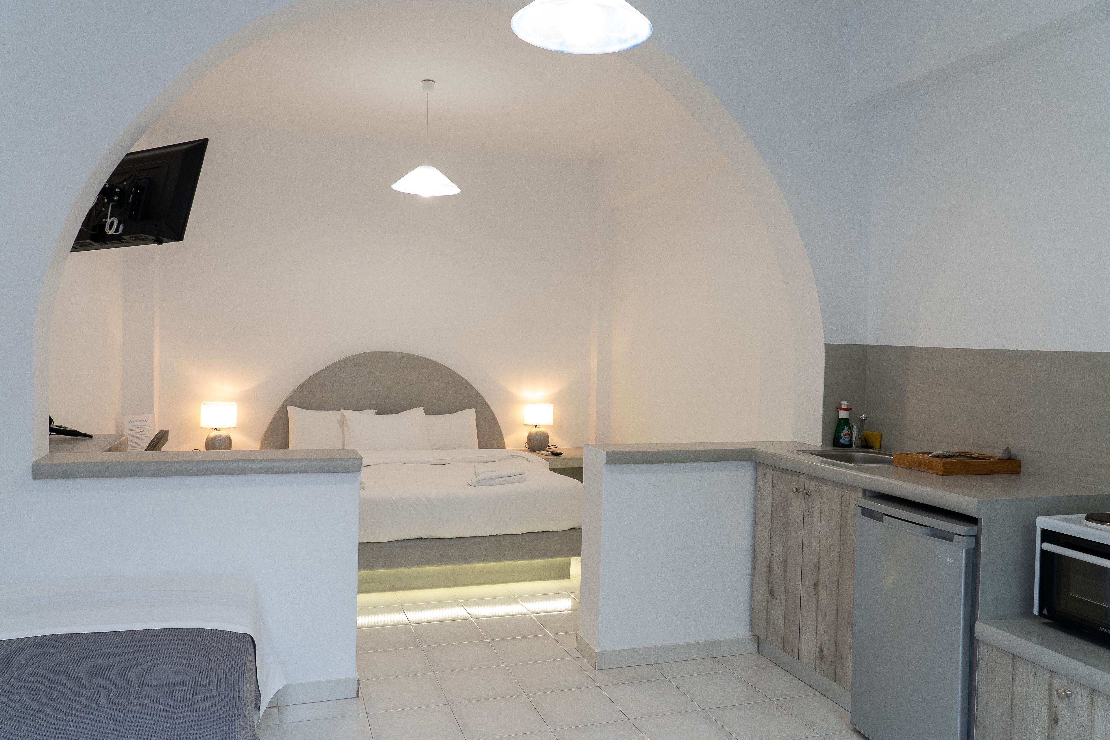 Quarto de hotel moderno com cama, mesa de cabeceira, luminária e um espaço com cozinha compacta.