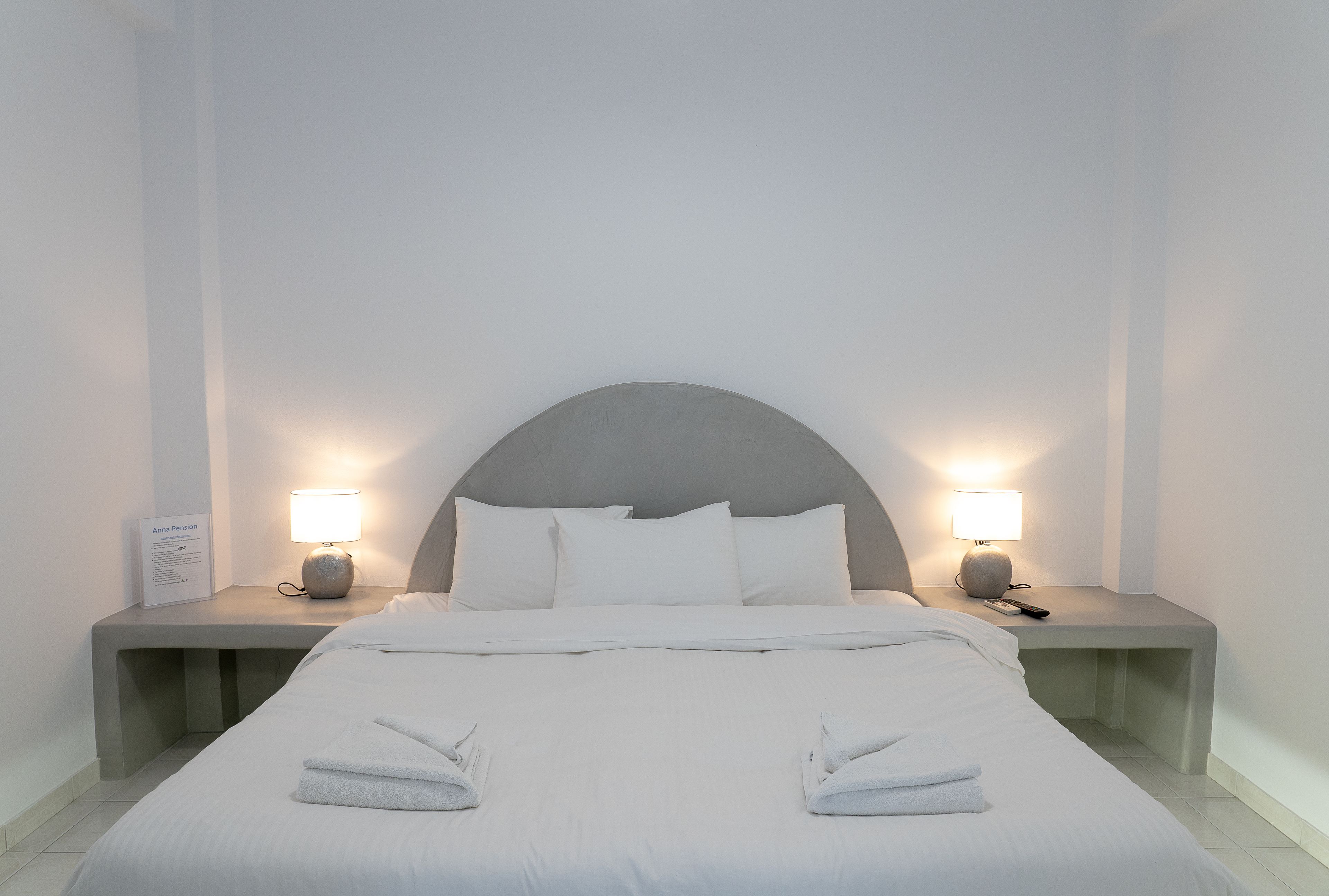 Una cama bien hecha con almohadas y una toalla doblada, dos mesitas de noche con lámparas, y un cartel en la mesa de la izquierda.