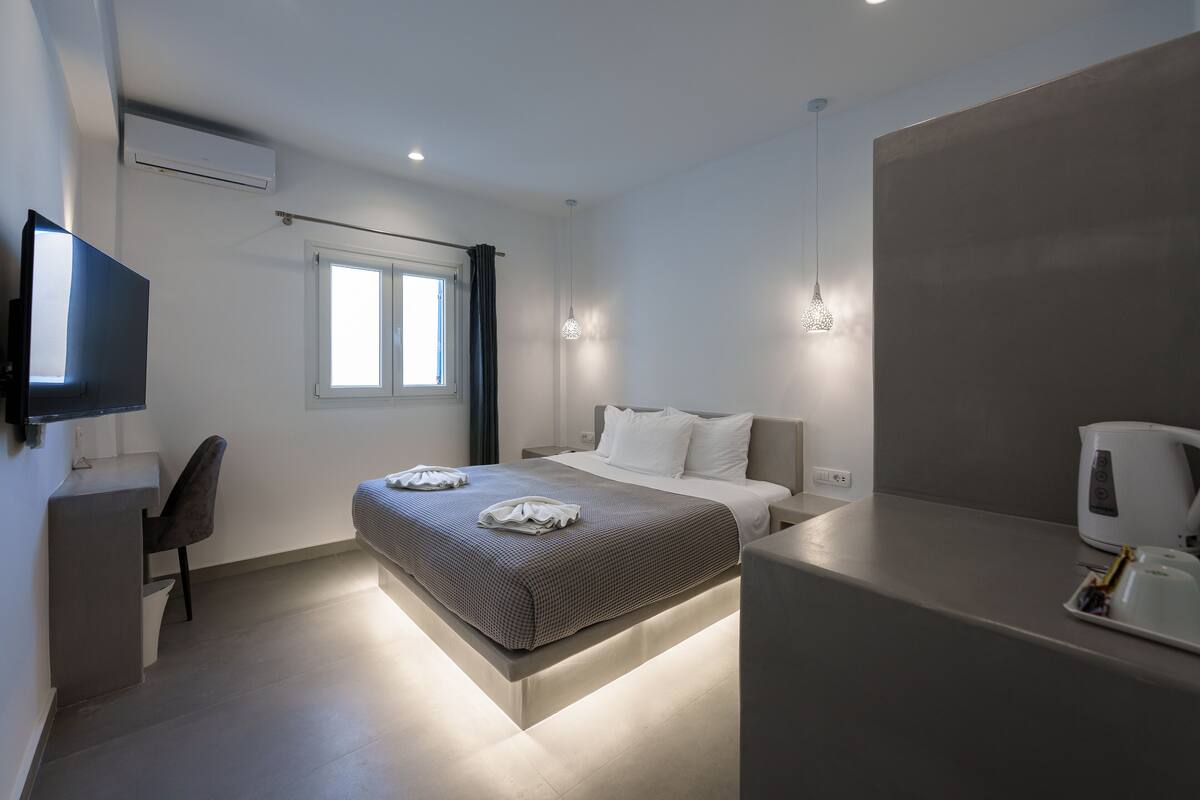Una habitación de hotel moderna con cama, mesita de noche, televisor y ventana con cortinas.
