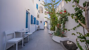 Un callejón estrecho con paredes blancas y contraventanas azules, que conduce a un edificio con una entrada arqueada. Hay sillas y mesas blancas a lo largo del camino, y una planta con flores rosas a la derecha.