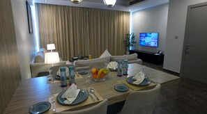 A dining area with a long table set for four, a sofa, a TV, and a plant.
