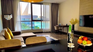 Une chambre d’hôtel moderne dotée d’une grande fenêtre donnant sur une piscine et des bâtiments, d’un canapé avec des coussins jaunes, d’une table à manger avec des chaises, d’un téléviseur fixé au mur et d’un panier de fruits posé sur la table.
