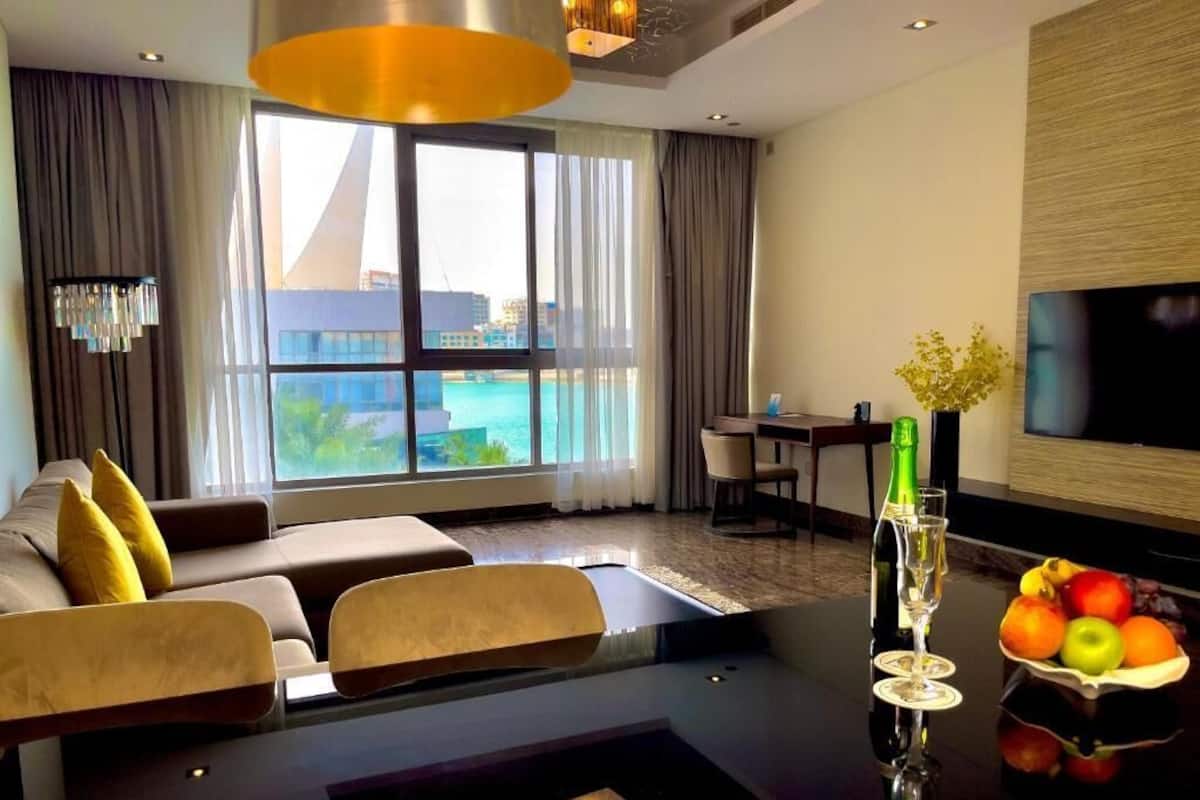 A modern hotel room with a large window offering a view of a pool and buildings, a sofa with yellow cushions, a dining table with chairs, a TV mounted on the wall, and a fruit bowl on the table.