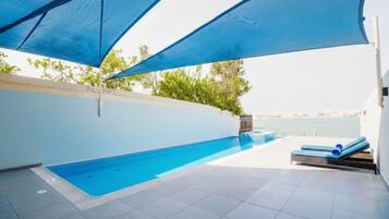Piscina externa com vista para o mar, área de estar com espreguiçadeiras e zona coberta com toldos azuis.