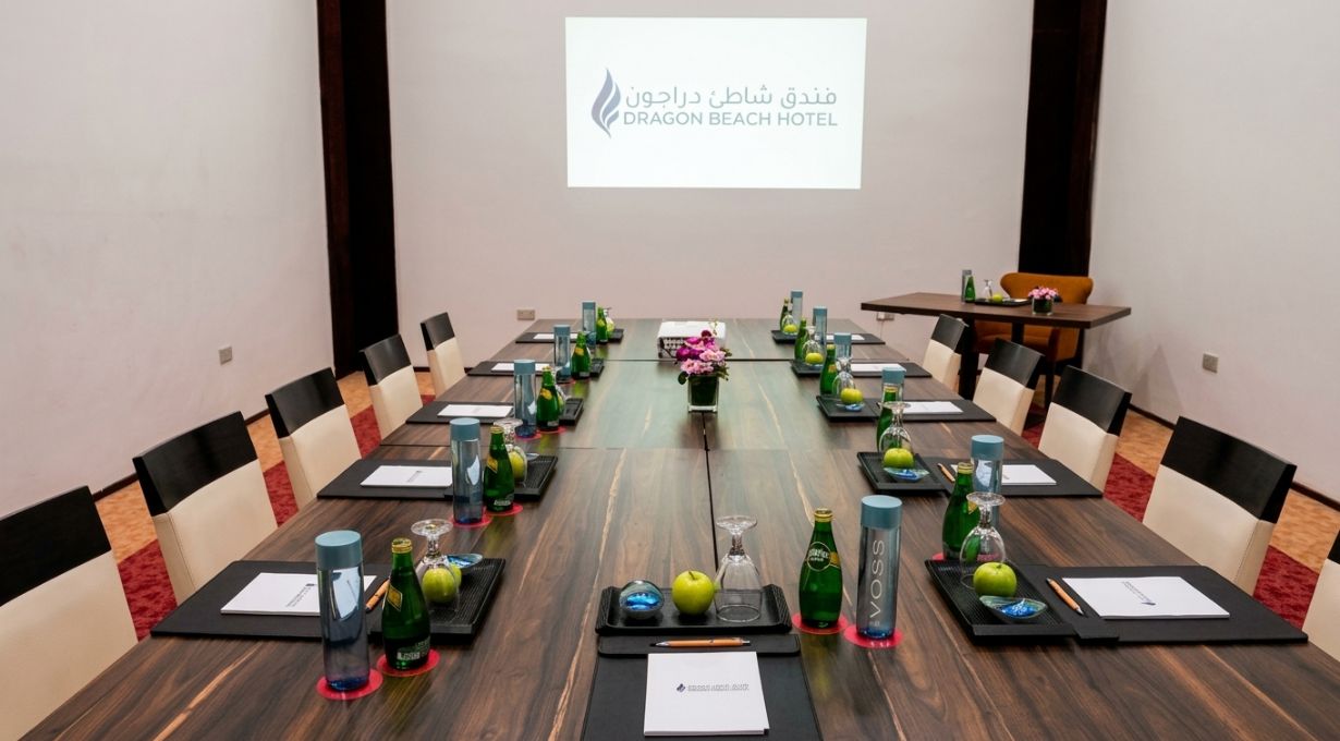 Un salón de conferencias con una larga mesa, sillas y una pantalla de proyector mostrando el texto 'Dragon Beach Hotel'.