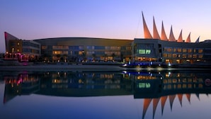 Un bâtiment moderne à la façade incurvée et aux nombreuses fenêtres, dont le reflet se mire dans une étendue d’eau calme.