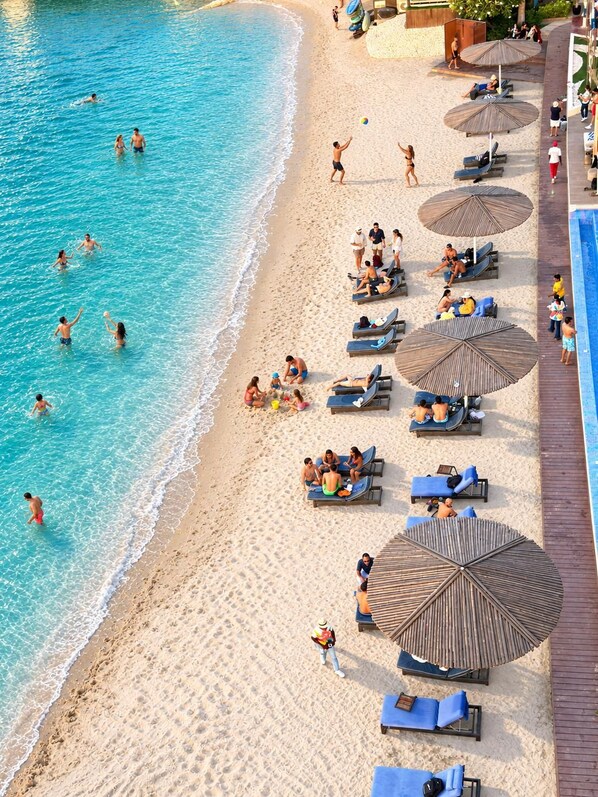 A beach with clear blue water, sun loungers, umbrellas, and people swimming and relaxing.