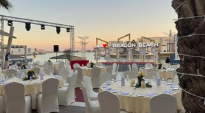 An outdoor dining area with round tables set for a meal, overlooking a body of water and a building with a balcony.