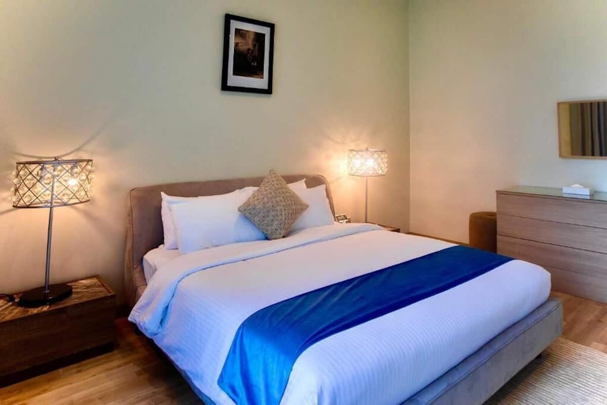 A neatly made bed with a blue accent, two bedside lamps, a framed picture on the wall, and a wooden dresser.