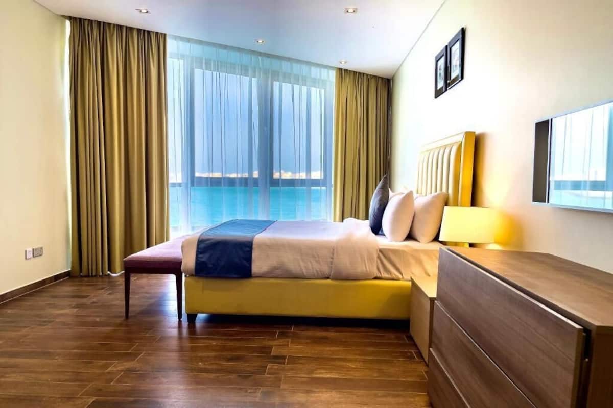 A hotel room with a large bed, a bedside table, a lamp, and a view of a pool through sheer curtains.