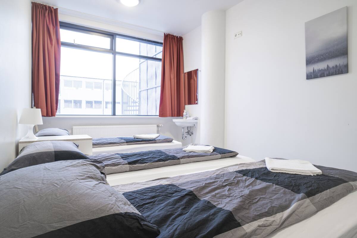 A hotel room with two beds, a window with curtains, a sink, and a painting on the wall.