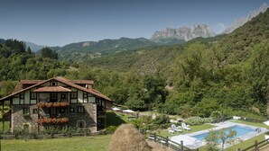 Un chalet à la façade en pierre et au toit en pente, entouré d’une végétation luxuriante et de montagnes.