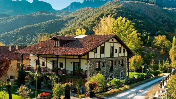 Une maison traditionnelle à colombages, dotée d’un balcon, entourée d’une végétation luxuriante et avec des montagnes en toile de fond.
