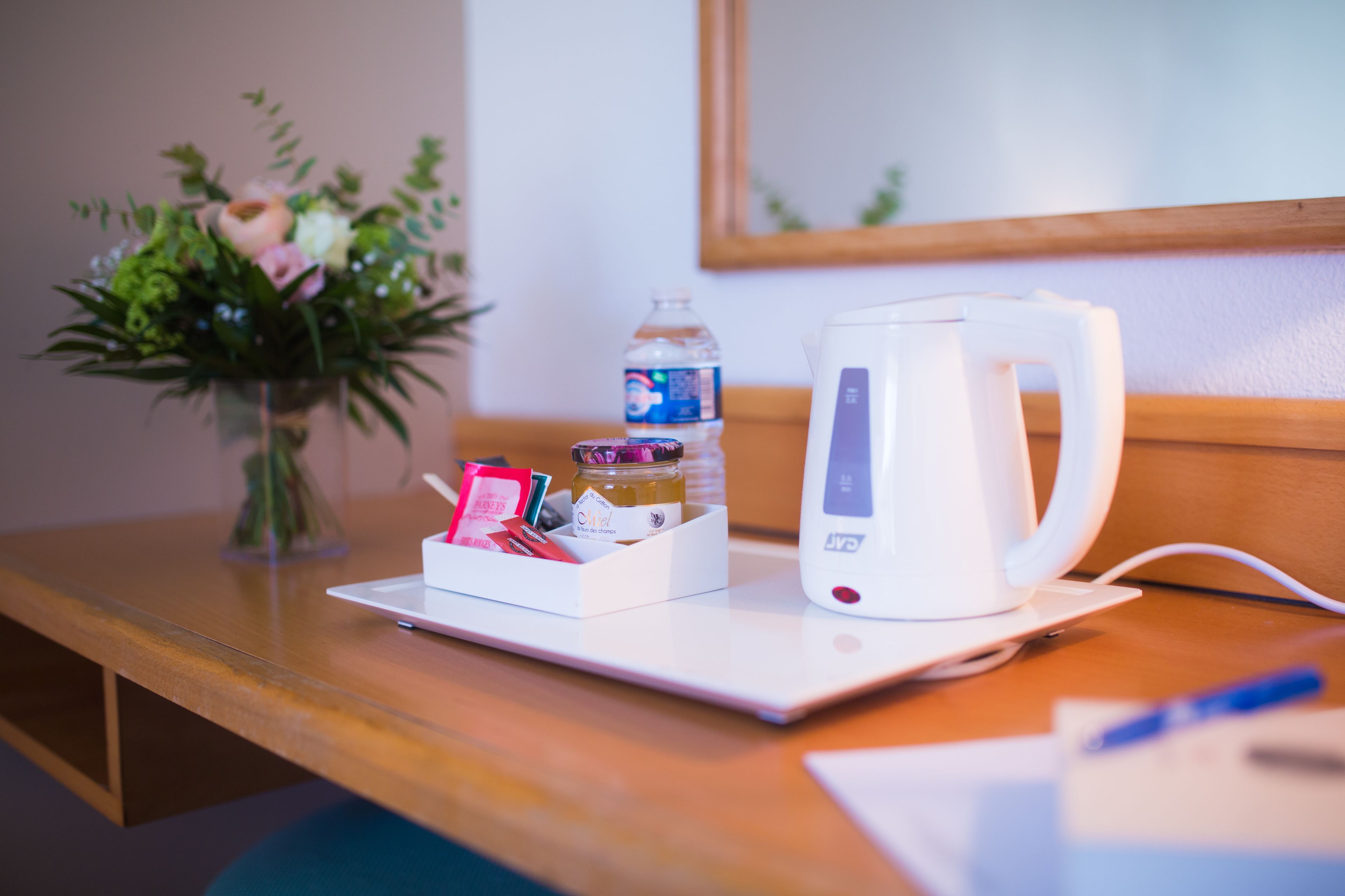 A wooden table with a white electric kettle, a water bottle, a small white box with items, and a vase with flowers.