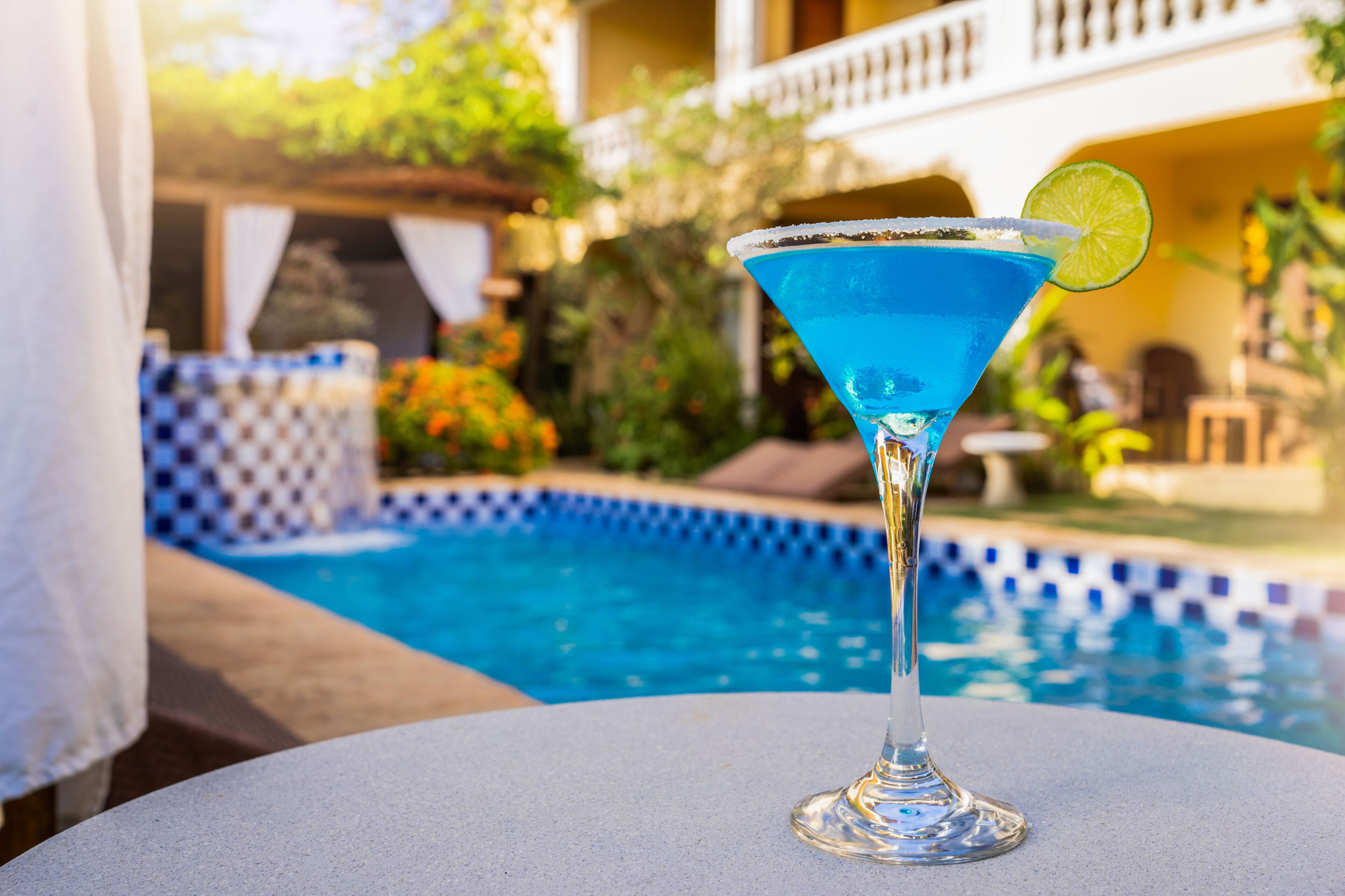 Una piscina azul cristalina con borde de azulejos, una mesa blanca con un cóctel azul y un toque de lima.