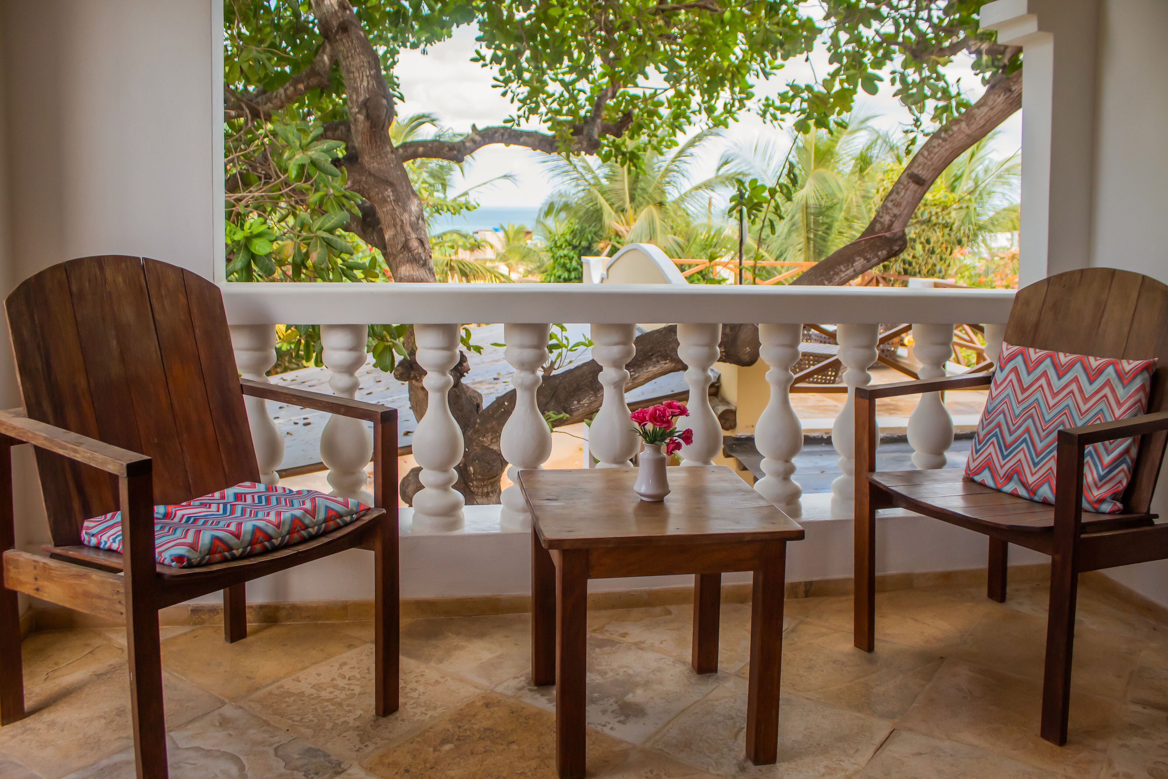 A balcony with two wooden chairs and a small table, overlooking a tropical landscape with trees and a view of the sea.