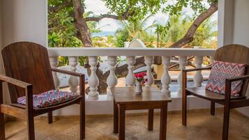A balcony with two wooden chairs and a small table, overlooking a tropical landscape with trees and a view of the sea.