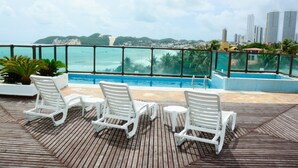 Un espace aménagé au bord de la piscine, avec des chaises longues blanches et une vue sur l’océan et le paysage urbain.