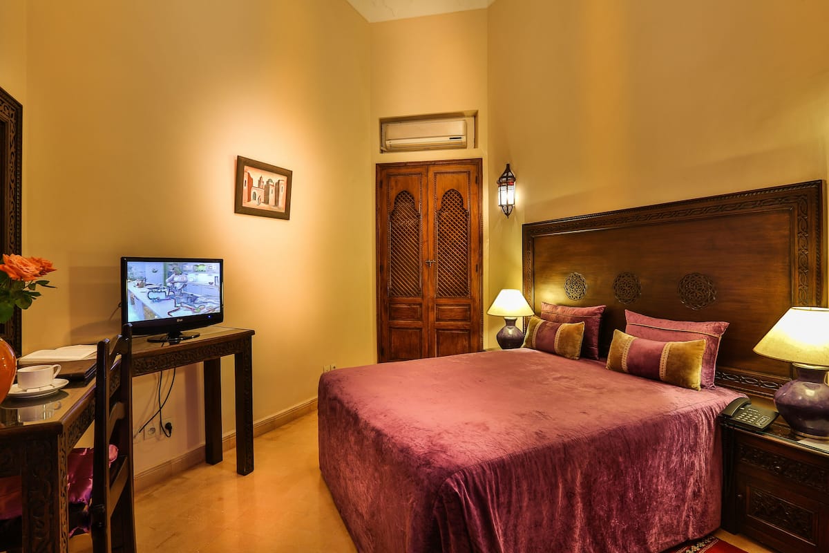 A hotel room with a large bed, a television on a small table, a desk with a chair, and a nightstand with a lamp.